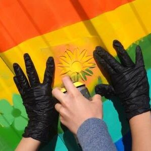 Zwei Hände halten eine Schablone in Form einer Sonnenblume an die Wand, damit eine dritte Hand durch Schablone Farbe sprühen kann.